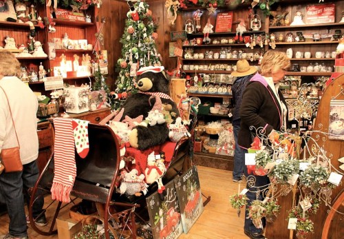 A cozy holiday shop filled with decorations, a bear in a sleigh, and shoppers browsing festive items.