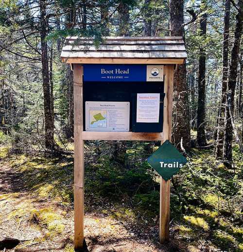 A wooden trailhead sign in a forest, displaying "Boot Head" and a map, with a green "Trails" marker below.