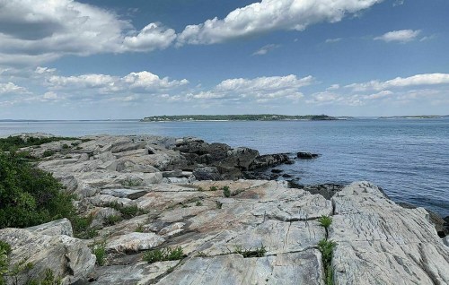 A rocky shoreline with calm water and a distant island under a blue sky with fluffy clouds.