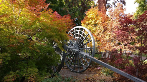 A large, metallic wheel structure surrounded by vibrant autumn foliage in a serene garden setting.