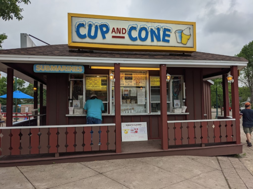 A small ice cream stand named "Cup and Cone" with a menu and customers outside on a cloudy day.