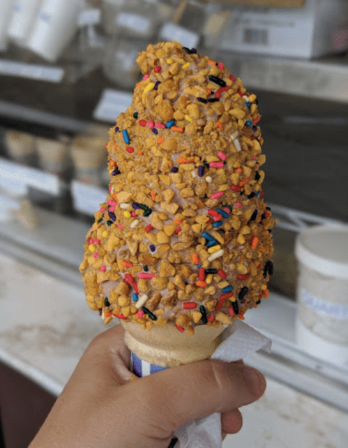 A hand holding a cone of ice cream topped with crushed peanuts and colorful sprinkles.