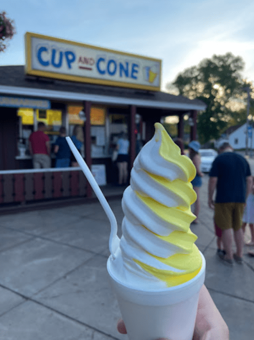 A hand holds a soft serve ice cream cone with yellow swirl, in front of a shop called "Cup and Cone."