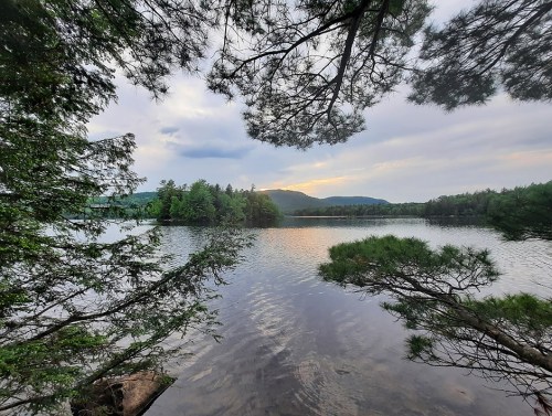 A serene lake surrounded by trees, with mountains in the background and a cloudy sky reflecting on the water.