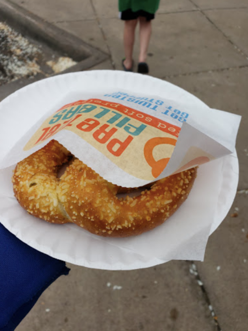 A hand holding a sesame seed pretzel on a paper plate, with a food wrapper partially covering it.