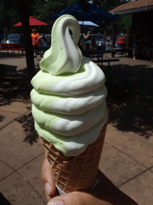 A hand holding a swirl of green and white soft serve ice cream in a waffle cone, with outdoor seating in the background.