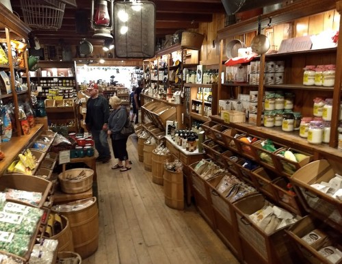 A vintage grocery store interior with wooden shelves, barrels, and customers browsing various products.