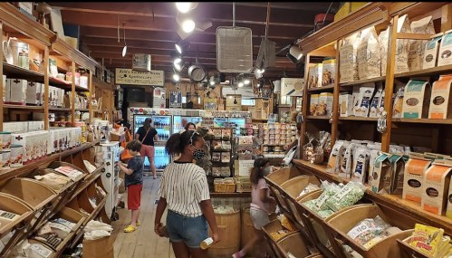 A bustling store interior filled with shelves of packaged goods, shoppers browsing, and a wooden display in the center.