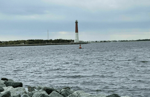 A lighthouse stands on the shore, with a buoy in the foreground and calm waters under a cloudy sky.
