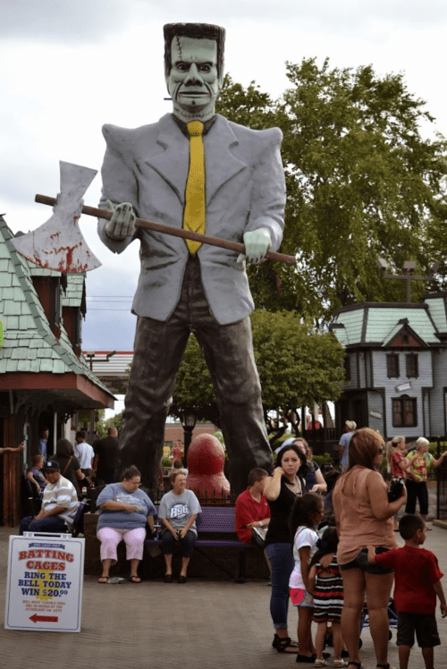 A large statue of a man in a suit holding an axe stands in an amusement park, with visitors gathered around.
