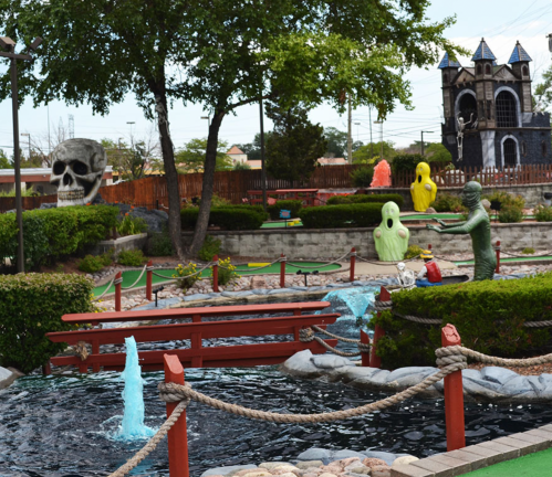 A mini-golf course featuring spooky decorations, including a skull, ghosts, and a castle, surrounded by greenery and water features.