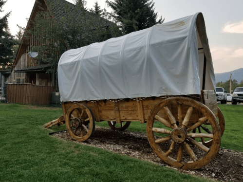 A wooden covered wagon on display in a grassy area, with a rustic building and trees in the background.