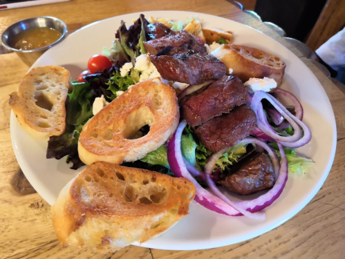 A plate of grilled meat served on a bed of mixed greens, red onions, and toasted bread, with a small bowl of sauce.