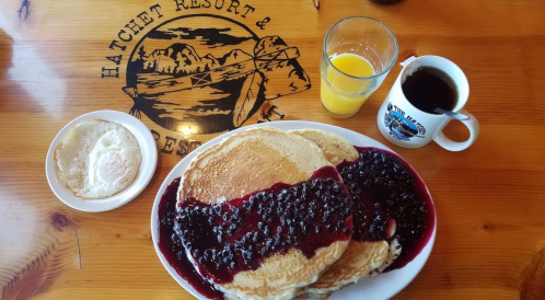 A plate of pancakes topped with berry sauce, alongside a glass of orange juice and a cup of coffee on a wooden table.
