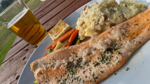 A plate with grilled salmon, mashed potatoes, carrots, and a slice of bread, alongside a glass of beer.