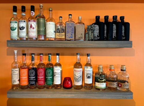 Two wooden shelves display a variety of liquor bottles against an orange wall, with a small red candle in the center.
