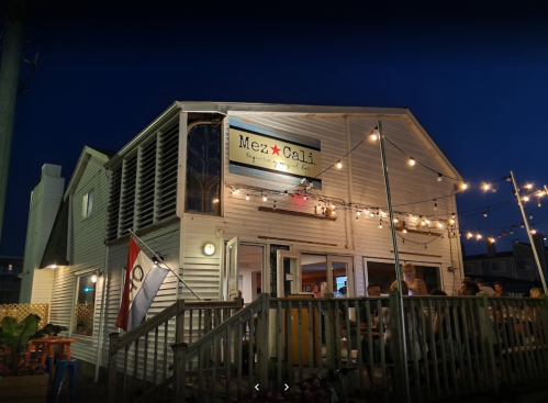 A cozy restaurant exterior at night, featuring string lights and a sign that reads "Mez Cali." People are dining outside.