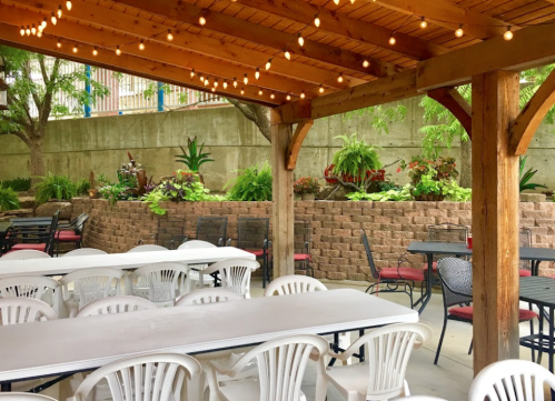 Outdoor dining area with wooden beams, string lights, and tables surrounded by greenery and a stone wall.