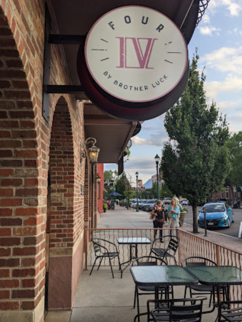 Sign for "Four by Brother Luck" outside a brick building, with outdoor seating and people in the background.