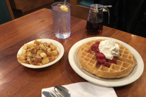 A plate with a waffle topped with strawberries and whipped cream, alongside a bowl of diced potatoes and a glass of water.