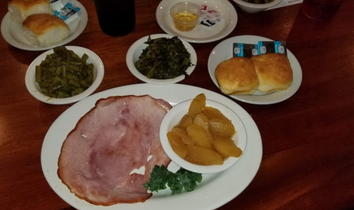 A plate with ham, greens, and peach slices, accompanied by rolls and sides of green beans and collard greens.