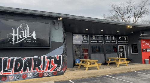 Exterior of a shop with a sign reading "Hail Dark Aesthetics," featuring picnic tables and a mural saying "Solidarity."