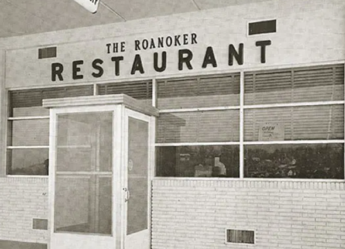 Black and white image of the Roanoker Restaurant exterior, featuring a glass door and large sign above.