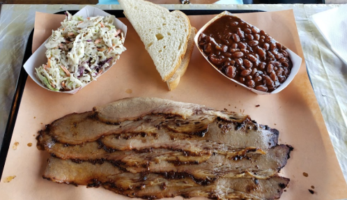 A plate featuring sliced brisket, coleslaw, baked beans, and a slice of bread on a brown paper surface.