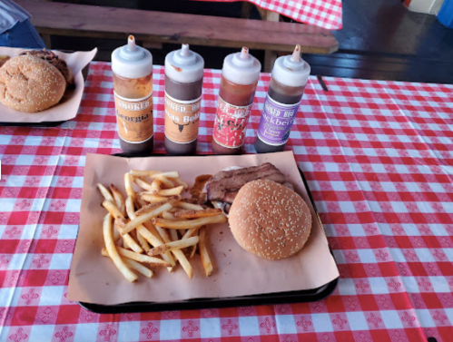 A plate with a burger, fries, and four bottles of barbecue sauce on a red-checkered tablecloth.