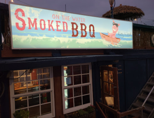 Sign for "On the Water Smoked BBQ" featuring a pig in a boat, illuminated against a twilight sky.