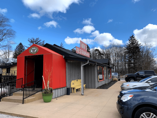 A red and gray restaurant building with a sign, surrounded by parked cars and trees under a blue sky with clouds.