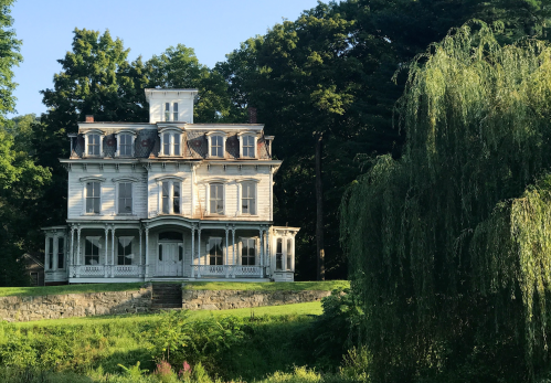 A large, historic white house with multiple stories and a wraparound porch, surrounded by lush greenery.