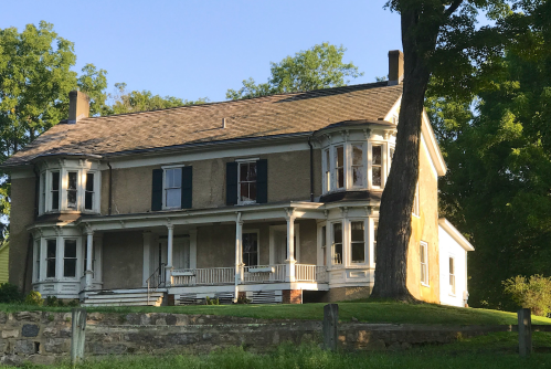 A historic two-story house with a wraparound porch, large windows, and a tree in the front yard, set against a clear sky.