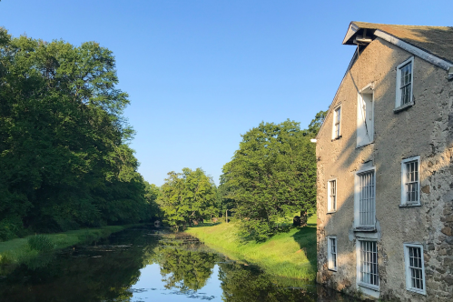 A serene canal scene with a stone building and lush greenery reflecting in the calm water under a clear blue sky.