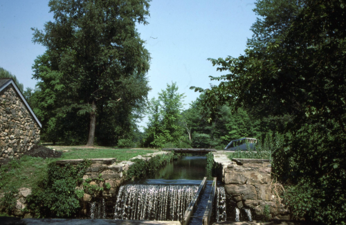 A serene landscape featuring a small waterfall, stone structure, and lush greenery under a clear blue sky.