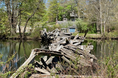 A dilapidated wooden bridge partially submerged in a calm river, surrounded by trees and greenery.