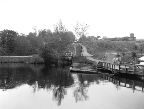 Black and white image of a calm river with trees, a wooden bridge, and a lock system in the background.