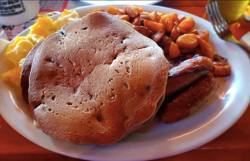 A plate of breakfast featuring a large pancake, scrambled eggs, sausage, and golden-brown potatoes.