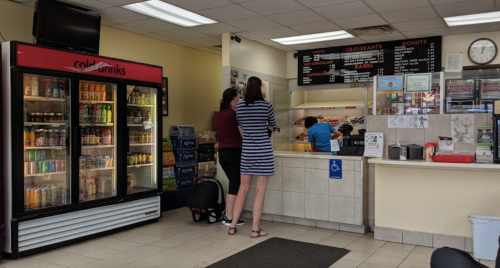 A café interior with a drink fridge, two customers at the counter, and a menu board on the wall.