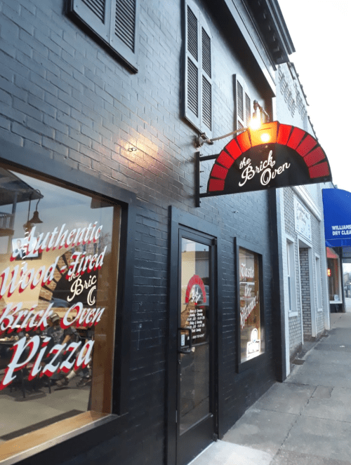 Exterior of The Brick Oven restaurant, featuring a black facade and a red awning with "Authentic Wood-Fired Pizza" sign.