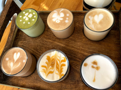 A wooden tray holds six cups of coffee with intricate latte art designs in various colors and patterns.