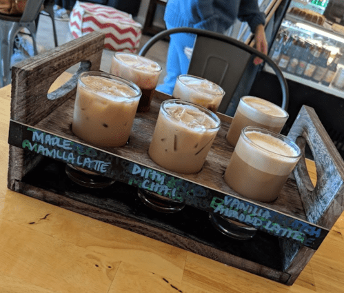A wooden tray holds eight glasses of various iced lattes, labeled with flavors like vanilla and chai.
