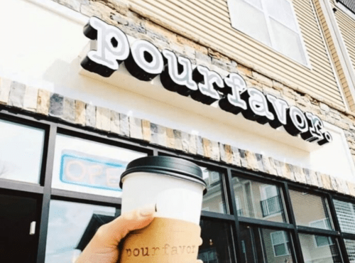 A hand holds a coffee cup in front of the "pourfavor" café sign, with an "OPEN" sign visible in the window.