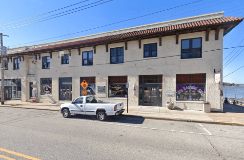 A two-story building with shops on the ground floor, located along a street by a river, under a clear blue sky.