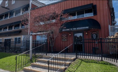 Exterior of a steakhouse with a welcoming sign, featuring a red building, black awnings, and a landscaped entrance.