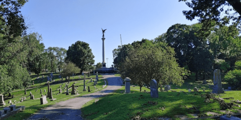 A peaceful cemetery scene with a tall monument and trees lining a winding path under a clear blue sky.