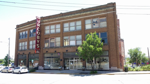 Historic brick building with large windows and a sign reading "Jacoby's," surrounded by trees and parked cars.