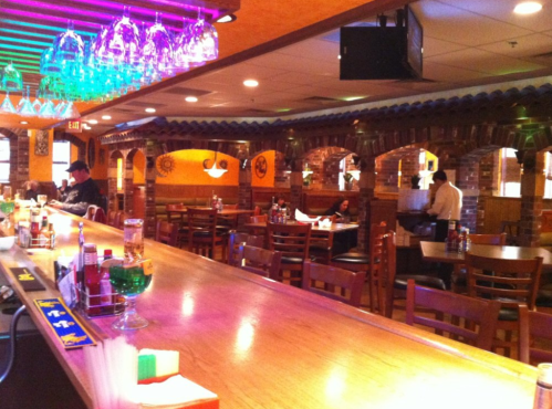 A vibrant bar interior with colorful lighting, wooden chairs, and a long counter, featuring patrons and a bartender.