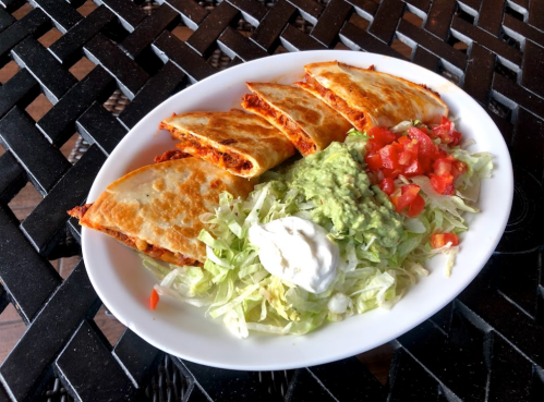 A plate of quesadillas with guacamole, salsa, lettuce, and sour cream, served on a woven table.