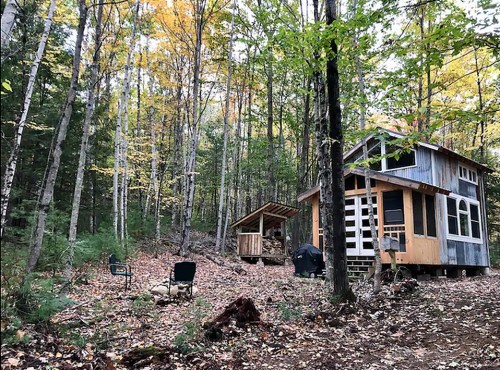 A cozy cabin in a forest, surrounded by autumn foliage, with a fire pit and seating area nearby.